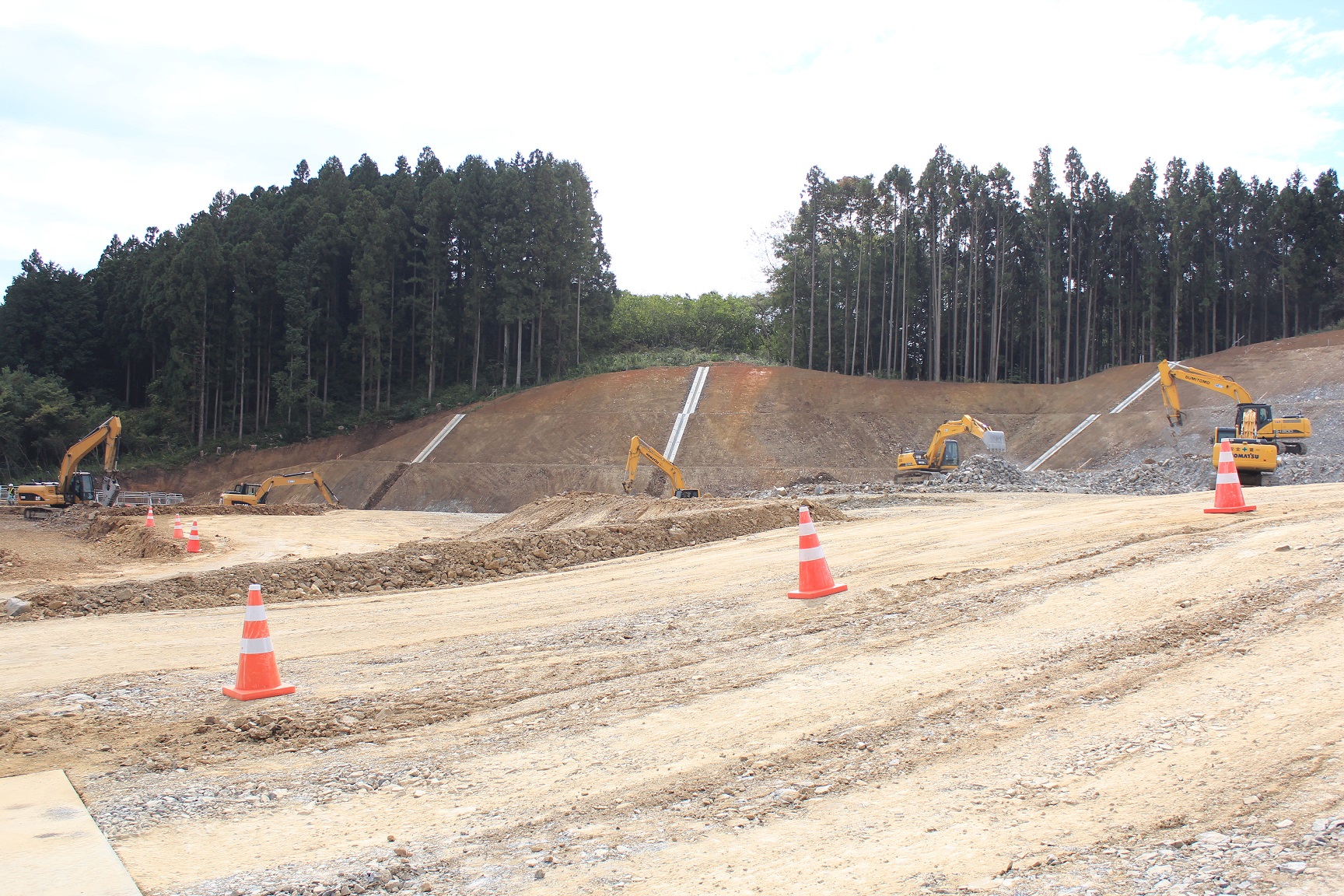 森林のふもとの斜面に沿って複数の重機が作業している造成地の写真
