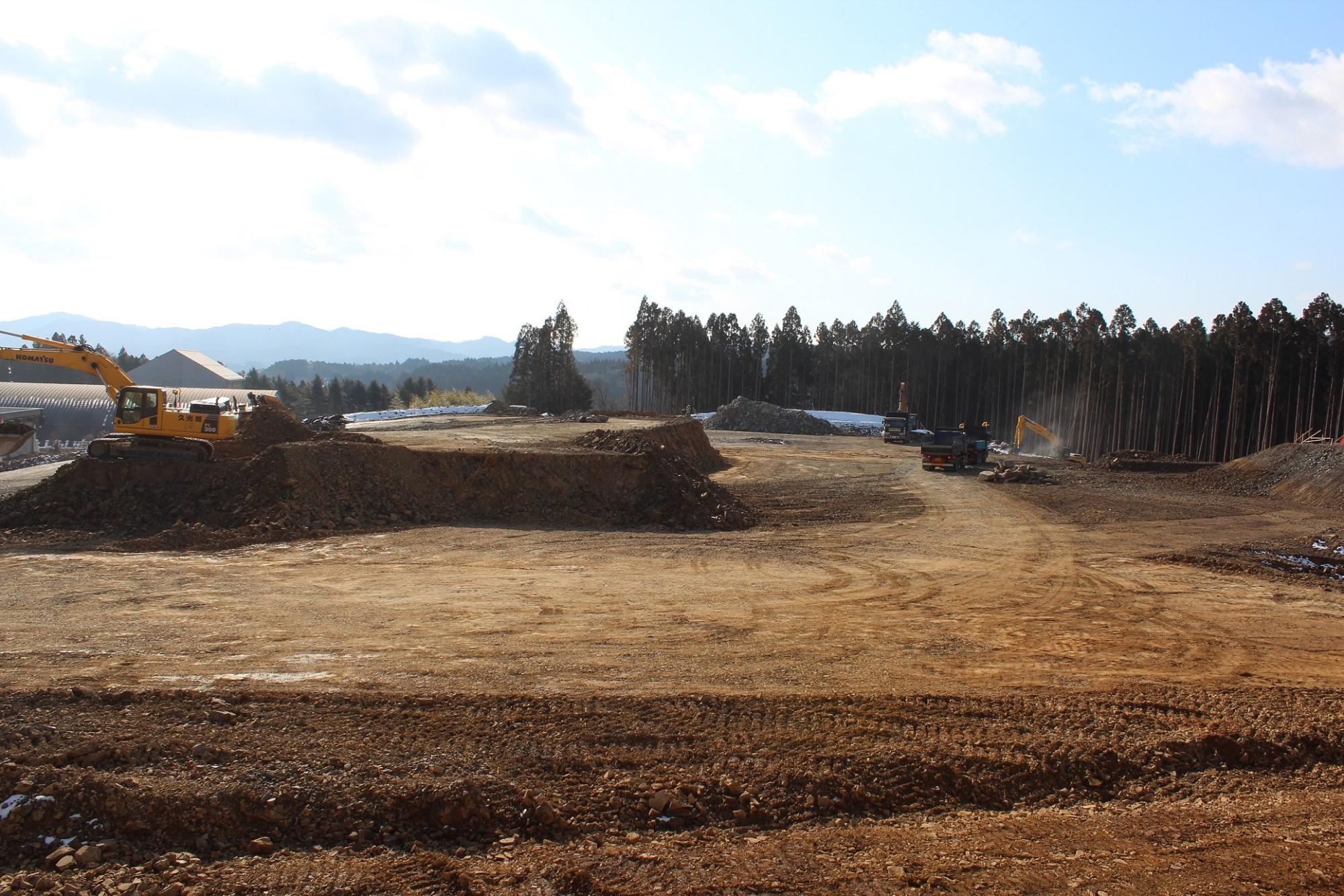 遠くの山々と林を背景に、重機が土砂を運ぶ広大な造成地の写真