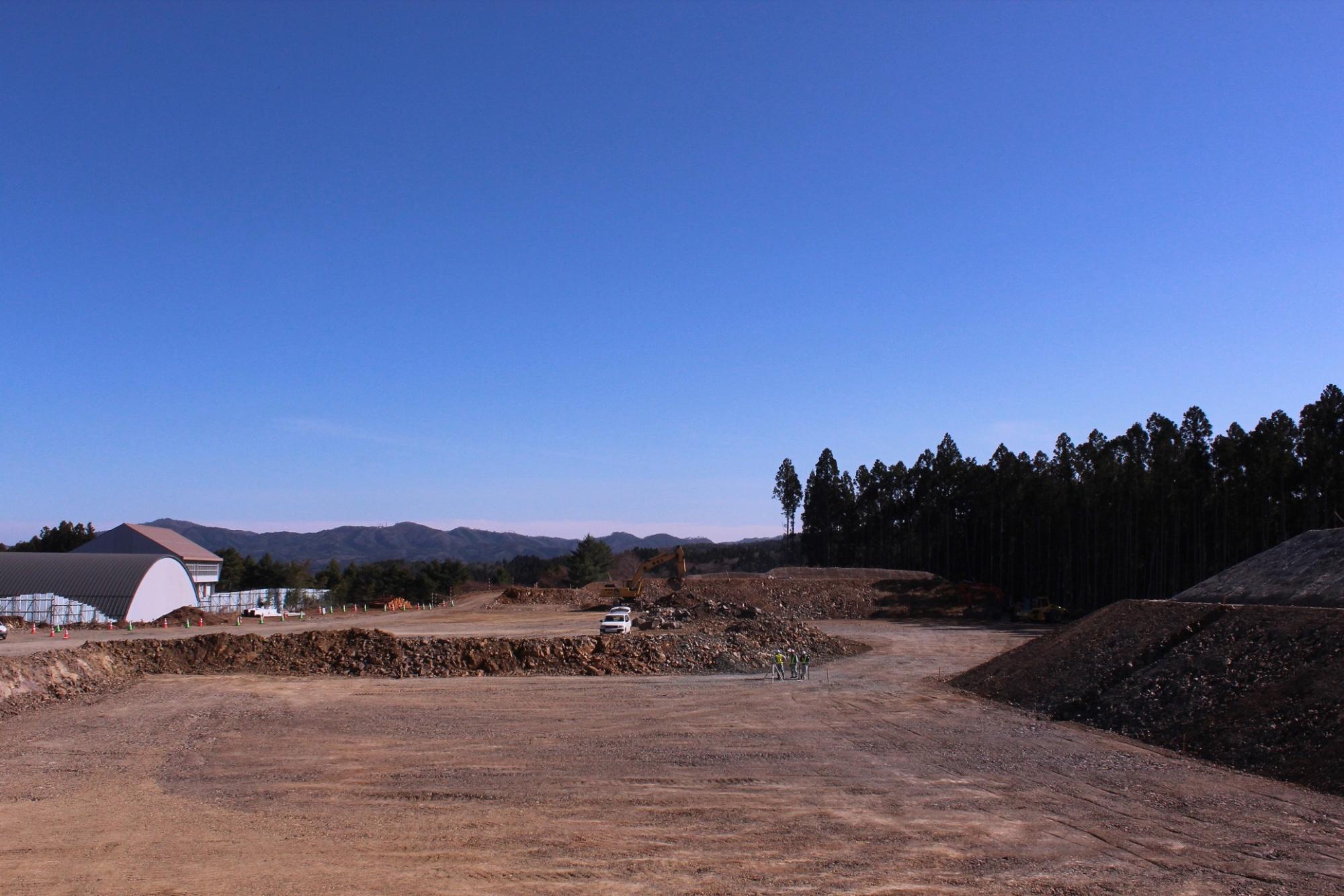 建物や遠くの山を背景に、広大な造成地で重機が作業する様子の写真