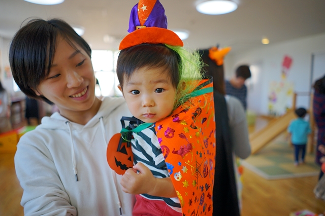 ハロウィンの室内イベントで母親がオレンジのマントと紫のとんがり帽子を身につけた幼い男の子を笑顔で抱きかかえている様子を写した写真