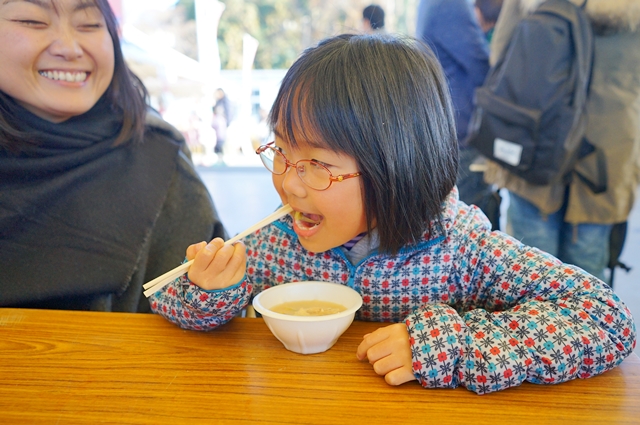 子供がテーブルでお椀に入った食べ物を箸で食べ、その様子を満面の笑みを浮かべた母親が見ている写真