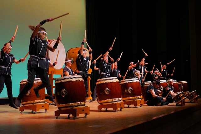 舞台の上で、複数の和太鼓奏者たちが黒い衣装と鉢巻きを身に着け、力強く太鼓を叩いたりジャンプしたりしながら躍動感あふれる演奏をしている様子の写真