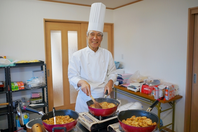 白いコック帽とコックコートを着た高橋恒雄さんが、室内で3つのフライパンを使って炒め物を調理しながら笑顔を見せている写真