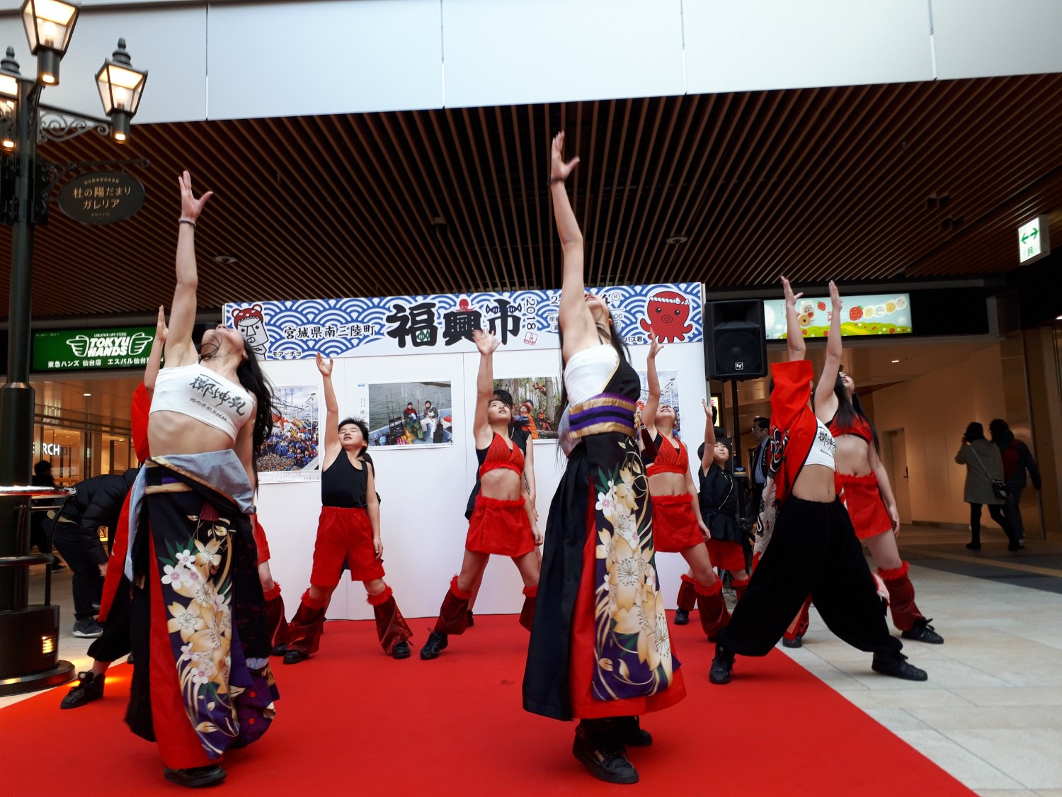 南三陸町の福興市のステージ上で、白いトップスに黒地の花柄袴風ボトムスや赤いショートパンツ、黒いパンツに赤い上着など赤と黒を基調とした衣装を身に着けた子どもたちと女性が、手を高く伸ばして力強い踊りを披露している写真