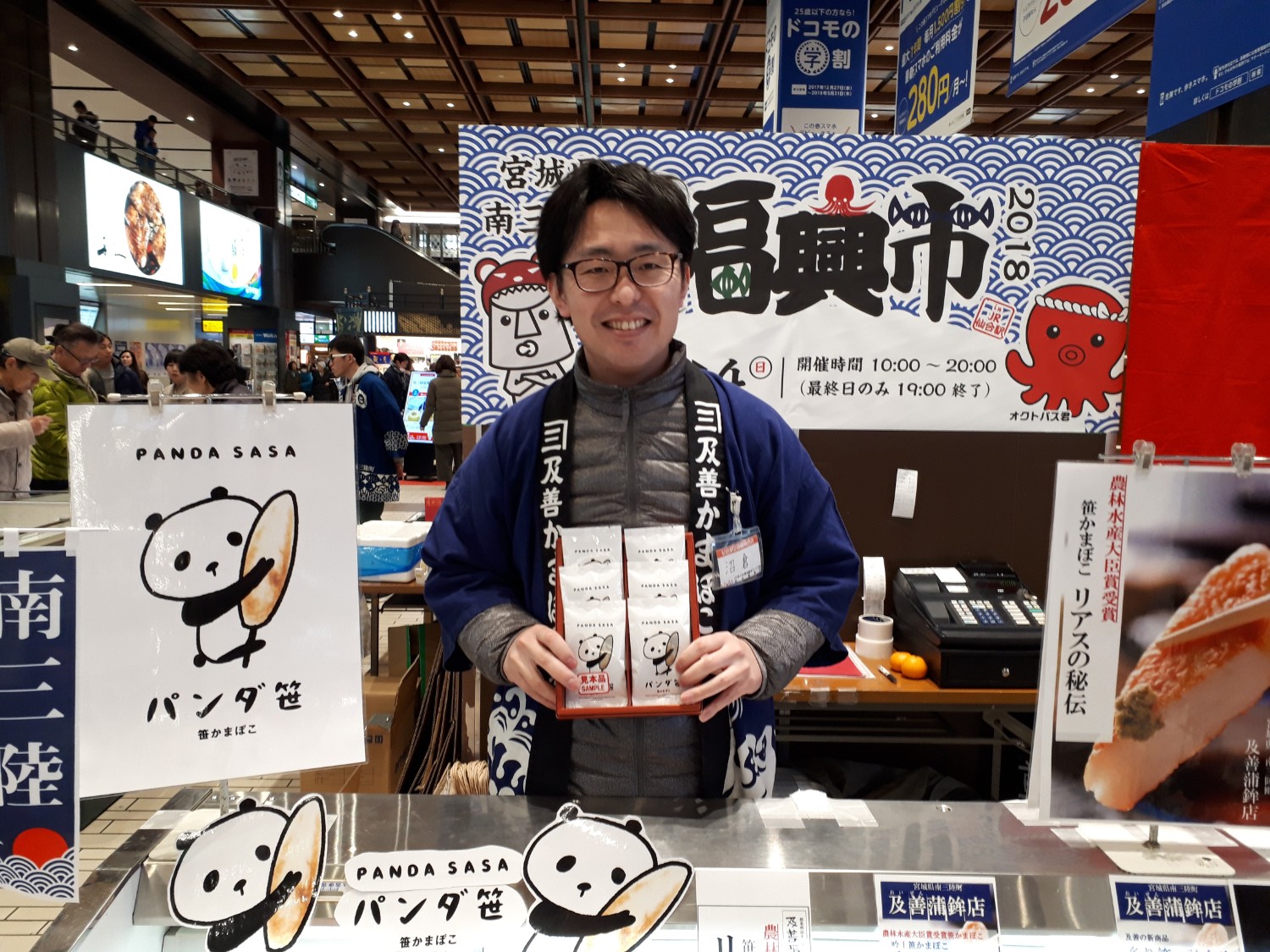 “宮城県 南三陸 福興市 2018”と書かれた装飾パネルを背景に、『パンダ笹（PANDA SASA）』という可愛いパンダイラストが描かれた笹かまぼこを笑顔で手に持つ、沼倉忠彦さんの写真