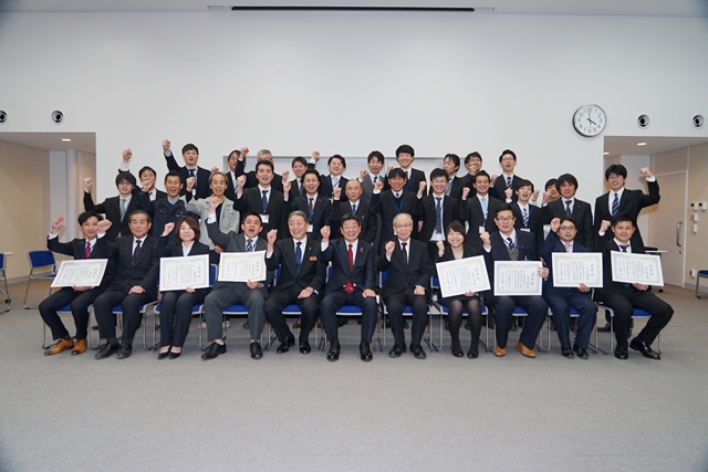 前列に4名の方と賞状を持った7名の方、後ろに整列して立っている多くの方々がガッツポーズをして記念撮影をしている写真