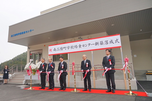 「祝 南三陸町学校給食センター新築落成式」と書かれた横断幕が掲げられた下で、佐藤町長を中心に4名の関係者がそれぞれハサミを持ちテープカットが行われようとしている写真