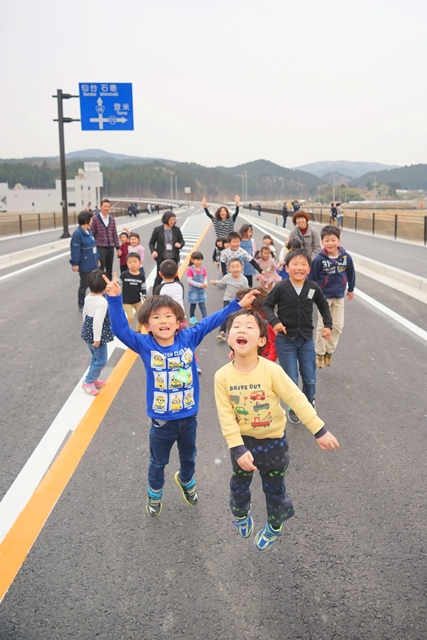 子どもたちと大人たちが新しくできた広い道路の上ではしゃぎながら歩いたり走ったりしていて、特に手前の男の子2人が笑顔でジャンプしているにぎやかで楽しげな様子を写した写真