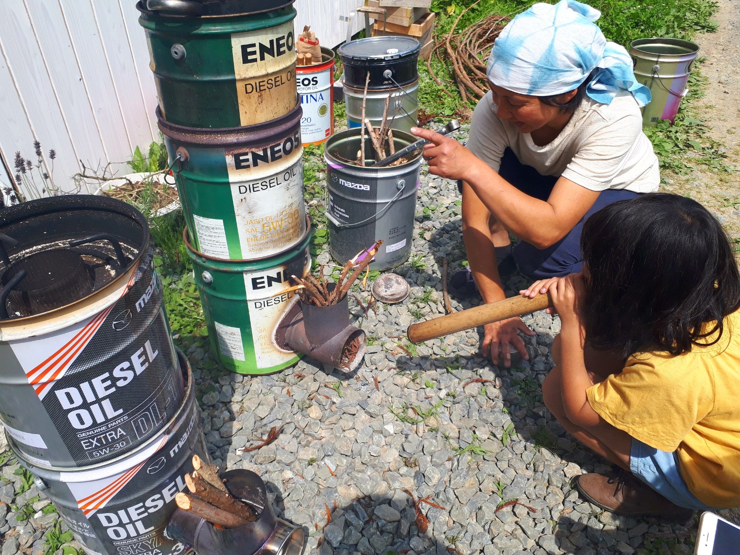 使用済みのペール缶を積み重ねたロケットストーブの前で、女性と子どもが地面にしゃがみながら火を起こしている様子の写真
