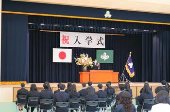 ステージ上には「祝入学式」の文字が掲げられ、紺色の制服を着た生徒達が並べられた椅子に座っている後姿を写した入学式の様子写真
