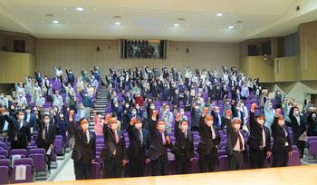 講堂の観客席で多くのスーツ姿の人々が立ち上がり、握りこぶしを作った右手を高く掲げている写真