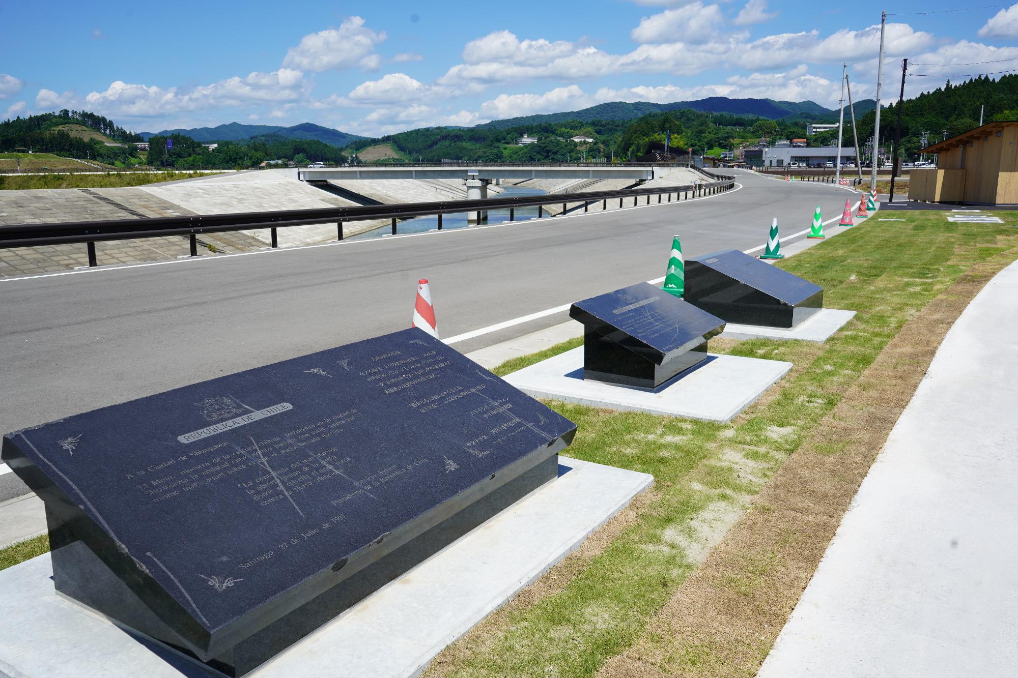 広々とした舗装された道路の脇に、3つの黒い石碑が芝生の上に設置されており、奥には山々と川、そして橋が見える風景の写真