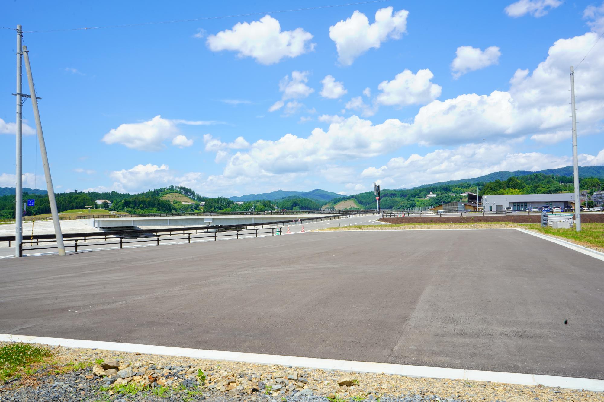 道路沿いにある舗装された広場の奥に山並みが見えるうみべの広場（B）の写真