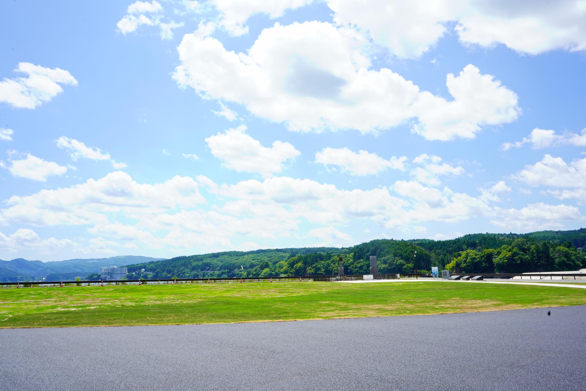 舗装された広場の隣に芝生の広場があり、その先には山や緑の森が広がっているうみべの広場（C）の写真