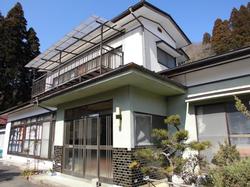 二階には屋根付きのベランダ、庭に植木があり、白と薄緑色の外壁に黒い瓦屋根を持つ二階建て住宅の外観写真