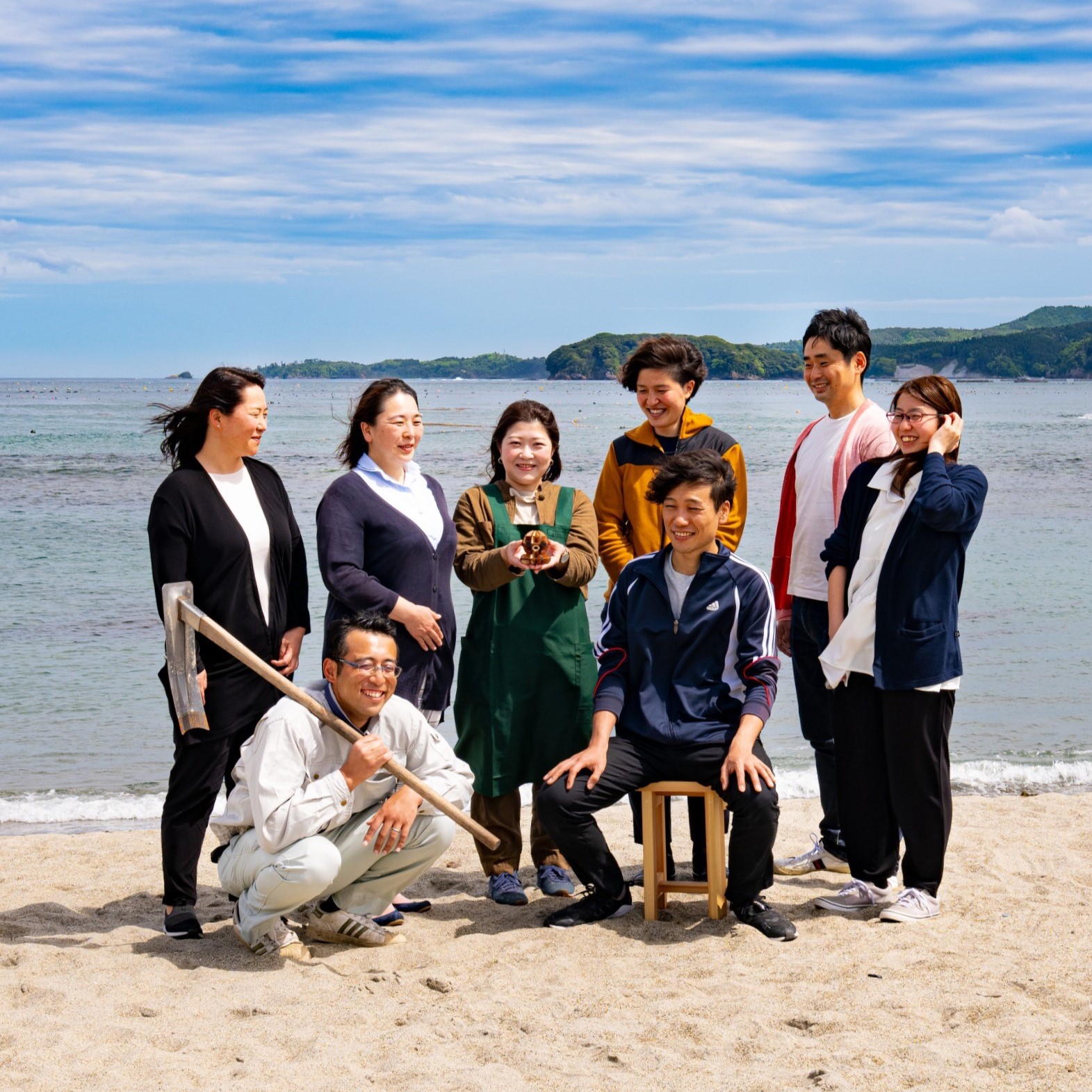 砂浜で海を背景に8人の男女が集まり、中央の男性が椅子に座り周囲の人々が立って笑顔を見せ、手前にはシャベルを担いでしゃがむ男性もいる、皆が和やかな雰囲気を漂わせている写真
