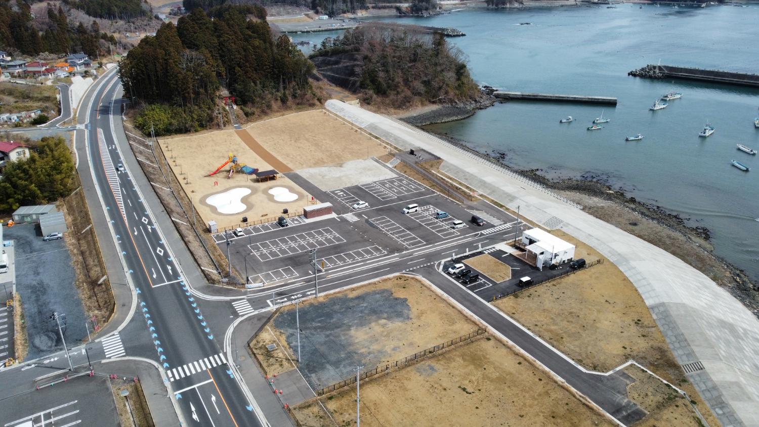 海に面した新しく整備された防潮堤と、その前に作られた公園と広い駐車場があり、園内にはカラフルな複合遊具などが整備されていて、背景には防波堤と海に浮かぶ漁船が見える昼間の風景を高台から定点カメラで撮影した写真