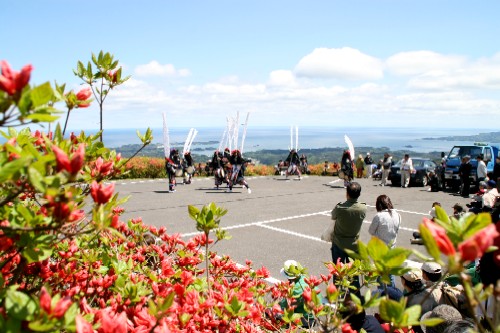 赤いツツジの花が咲いている山頂の駐車場のような広場で装束と面をつけ、背中に白い長い飾りを立てた人たちが伝統芸能を披露しており、観客がそれを鑑賞している様子の写真
