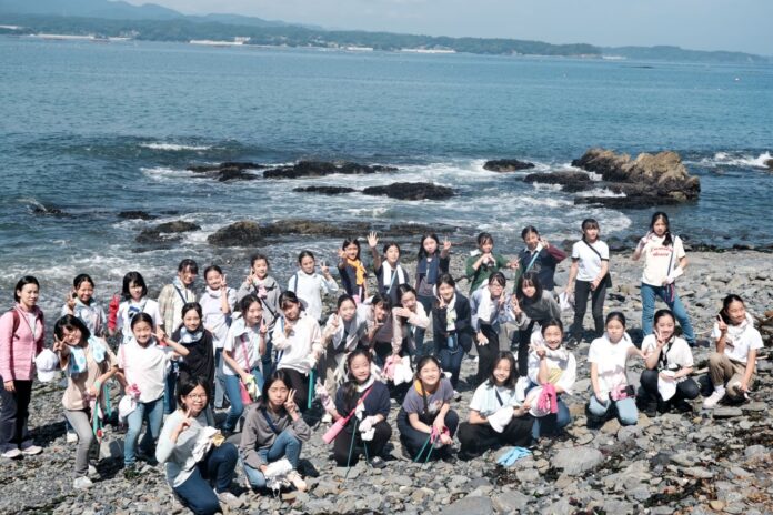 背景には波が打ち寄せる海が広がり、女子学生たちが海岸の岩場で清掃活動を行い、笑顔でポーズを取りながら記念撮影をしている写真