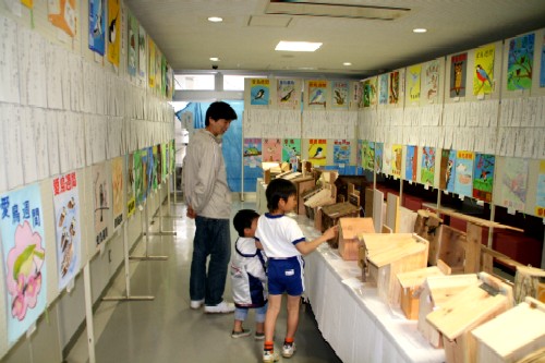 壁一面に鳥をテーマにした絵などが展示され、中央のテーブルには複数の手作りの巣箱が並んでいる場所に親子が作品を鑑賞している展示会の様子の写真