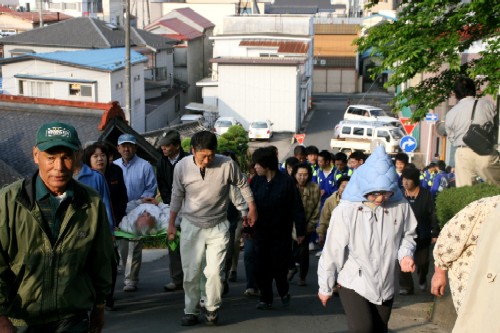 坂道を、複数の人たちが防災ずきんを被ったり簡易担架に横たわった人を運んだりしている訓練の様子の写真