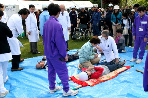 多くの見物人が集まる屋外の芝生の上で、青いビニールシートを敷き、負傷者役の二人が横たわり、白衣や救命胴衣を着用した医療関係者や救助隊員の方々が、応急処置などを行っている訓練の様子の写真