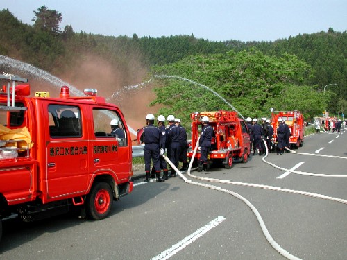 多くの消防服を着用した消防団員の方々が、道路上に並べられた数台の赤い消防車から白いホースを伸ばし、山に向かって勢いよく放水訓練を行っている様子の写真
