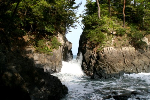 海辺の断崖絶壁が切り開かれた岩の間から、勢いよく白い波しぶきが打ち寄せており、その上部には濃い緑の木々が茂り、荒々しい岩と緑のコントラストが印象的な初夏の神割崎海岸の写真