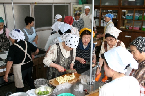 三角巾などを着けた複数の高齢の男女が、調理台で野菜や果物を切るなどの共同作業を行っているシルバークッキングの様子の写真