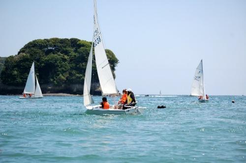 澄んだ青い海の上を、ライフジャケットを着用した数人の乗員を乗せた小型のヨットが、緑豊かな小島を背景にして進む様子を捉えた写真