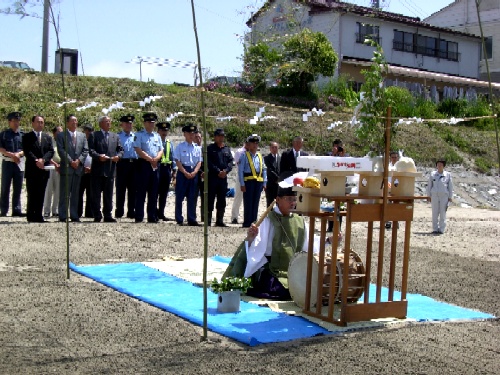 晴れた日に屋外の広い砂地で、青いビニールシートの上に座った神職が神事を行い、その後ろに背広姿と制服姿の多数の参列者が並んで見守る様子を捉えた写真