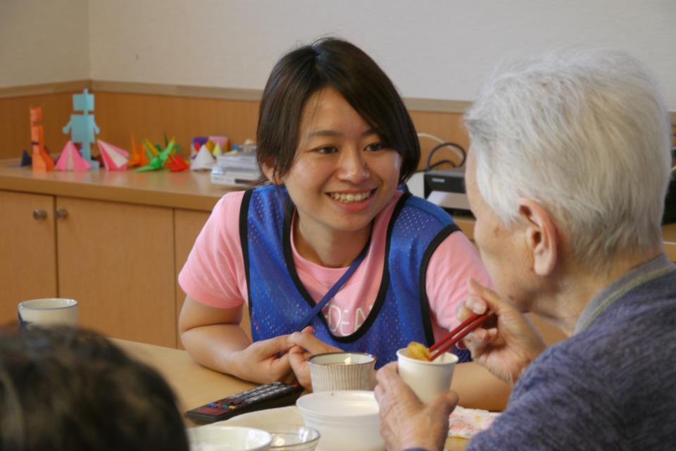 室内でピンク色のシャツと青いビブスを着た女性が、食事中の高齢の人と向き合いながら笑顔で話しかけている写真