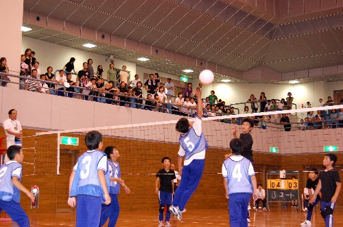 体育館の2階の観客席で多くの人が見守る中、青いビブスを着た生徒が空中で白いボールを打つ瞬間を捉えた、バレーボールの試合の様子の写真