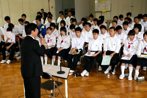 フローリングの床の広い部屋で、黒いスーツの男性がマイクで話をしており、制服姿の多数の生徒たちが並んで着席している様子を写した写真