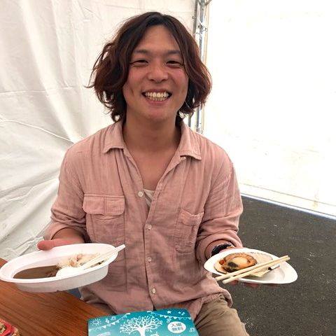 椅子に座り、両手にカレーライスと焼きホタテを持った笑顔の竹森さんの写真