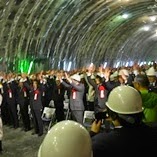 天井の照明で明るく照らされたトンネル内に椅子が列ごとに並べられ、ヘルメットを被りフォーマルな服装を着た大勢の人々が手を挙げている万歳三唱の様子の写真