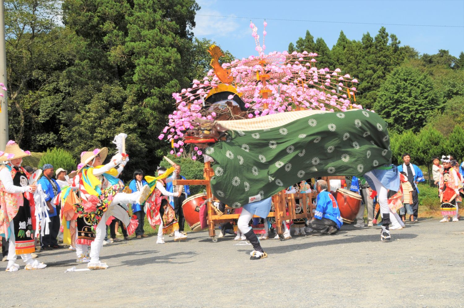 青空の下、しだれ桜の飾りがついた山車の前で、派手な衣装の踊り手と緑の布をかぶった獅子舞が躍動的に舞っている写真
