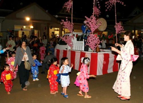 夜間の広場で、紅白幕や花飾りで装飾された軽トラックの荷台を舞台に見立てその上で太鼓を演奏している人と、その周りを浴衣姿の大人と子どもたちが楽しそうに踊っている様子の写真