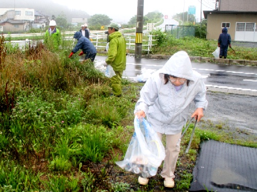 雨降る曇天の中、数人の人々が作業着姿で、道路脇の雑草やごみの除去作業に熱心に取り組んでいる様子の写真