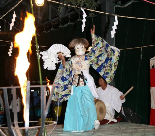 大きな炎が燃え盛る傍の舞台上で、青い袴と着物を着て白い面をつけた演者が、扇子を広げて両手を大きく広げて舞をおこなっている様子の写真