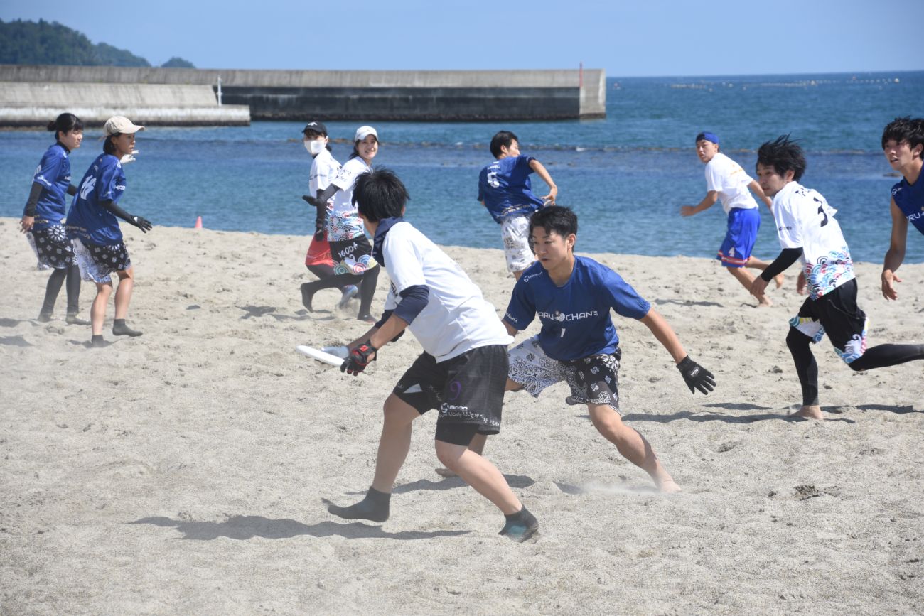 青と白のユニフォームを着た選手たちが砂浜でディスクを追いながら全力でプレーしている様子の写真