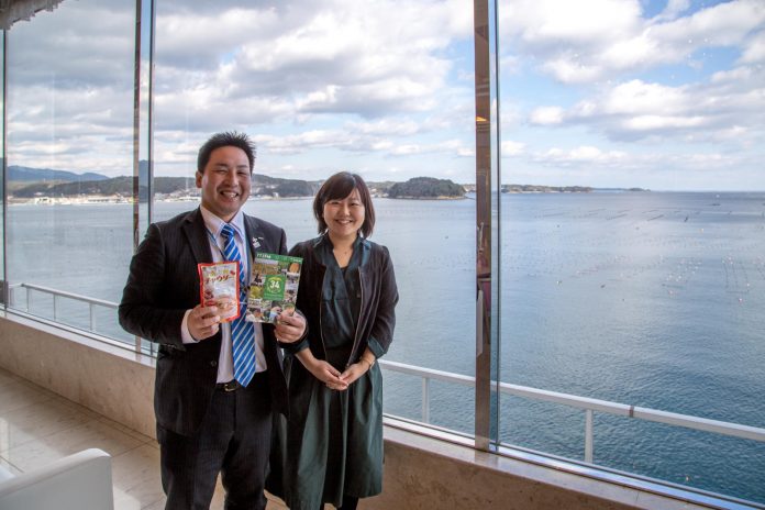 海を臨む大きなガラス窓の前に、女性リポーターと両手にチラシとチャウダーと書かれたレトルトパウチを持った伊藤さんが並んで立っている写真