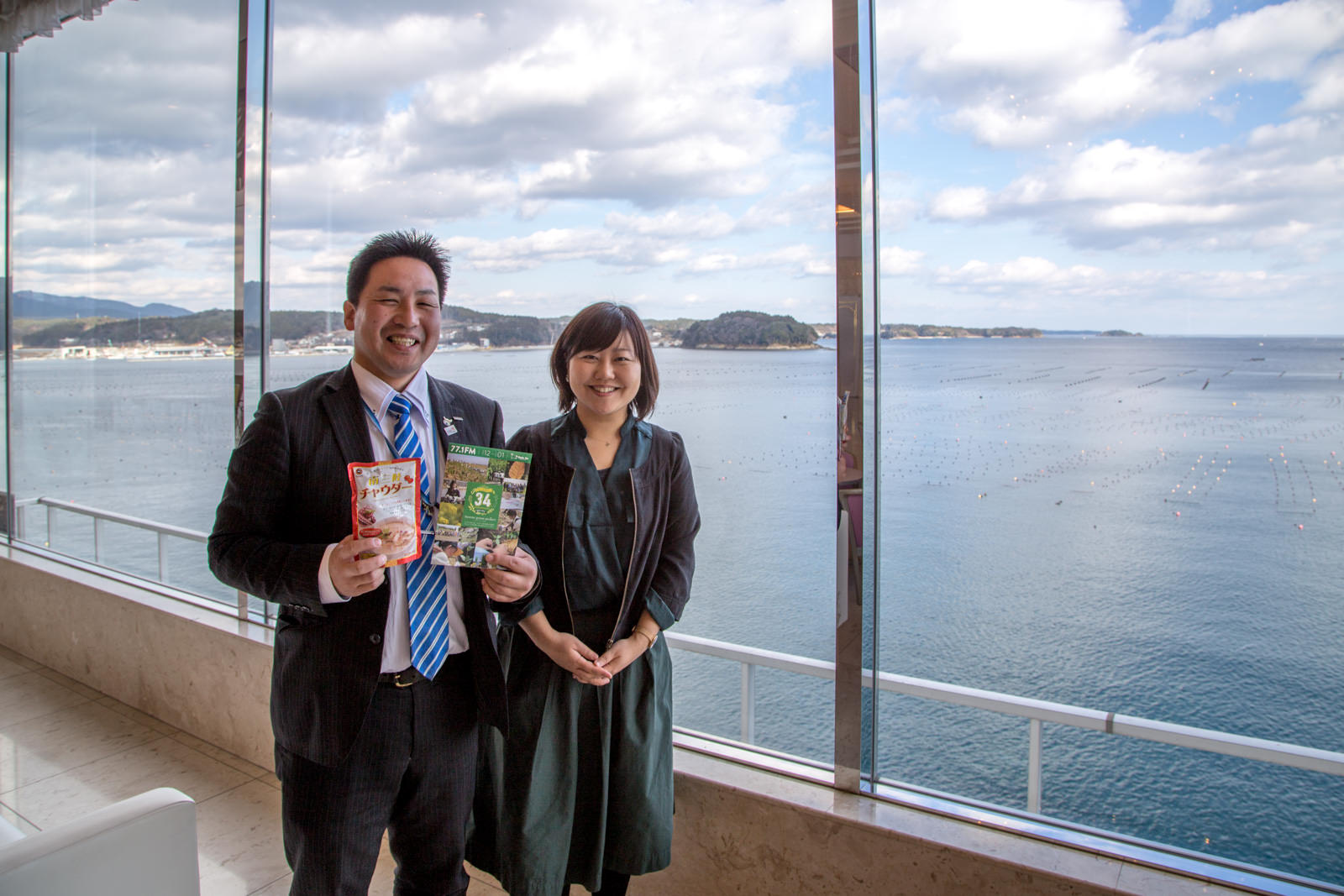 海を臨む大きなガラス窓の前に、女性リポーターと両手にチラシとチャウダーと書かれたレトルトパウチを持った伊藤さんが並んで立っている写真