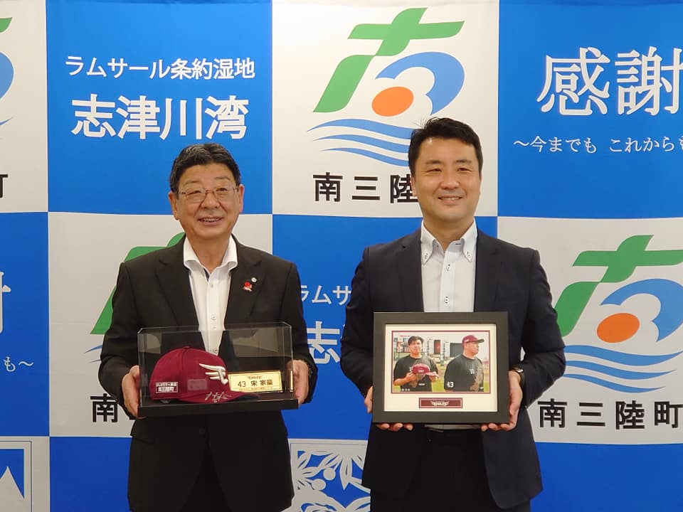 南三陸町の公式バックパネルの前で、佐藤町長が楽天イーグルスの帽子入りケース、代表の男性が記念写真の額縁をそれぞれ手に持ち、笑顔で並んでいる贈呈式の様子の写真