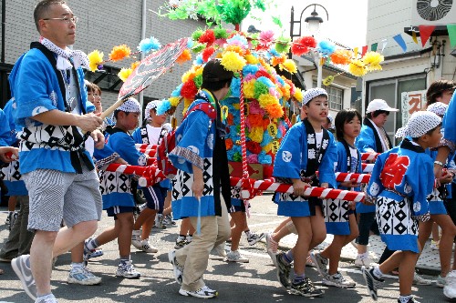 色鮮やかな花飾りで装飾された山車を、青い法被と手ぬぐいを身につけた大人と子どもたちが、元気よく担いで町中を練り歩いている様子の写真