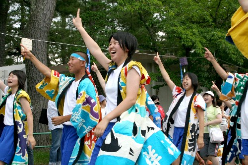 屋外で、お揃いのカラフルな衣装を着た男女が、満面の笑みで両手を天に突き上げながら、躍動感あふれる踊りを披露しているお祭りの写真