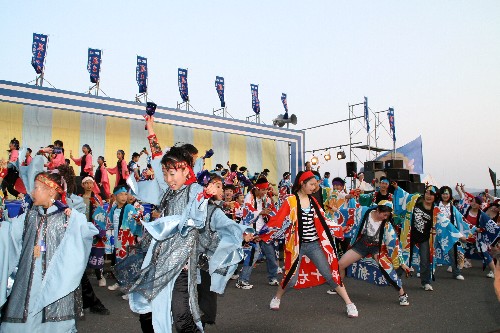 大勢の踊り手が、鮮やかな青や赤のお揃いの衣装を身につけ、活気あふれる表情で手を振り上げながらステージ前で熱心に踊りを披露しているイベント会場の様子の写真