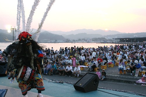 夕焼けの空と山々、海を背景に、広場に集まった大勢の観客の目の前で、鬼の面をつけ黒い髪と白い角、派手な衣装をまとった演者がステージ上で舞を披露している祭りの情景写真