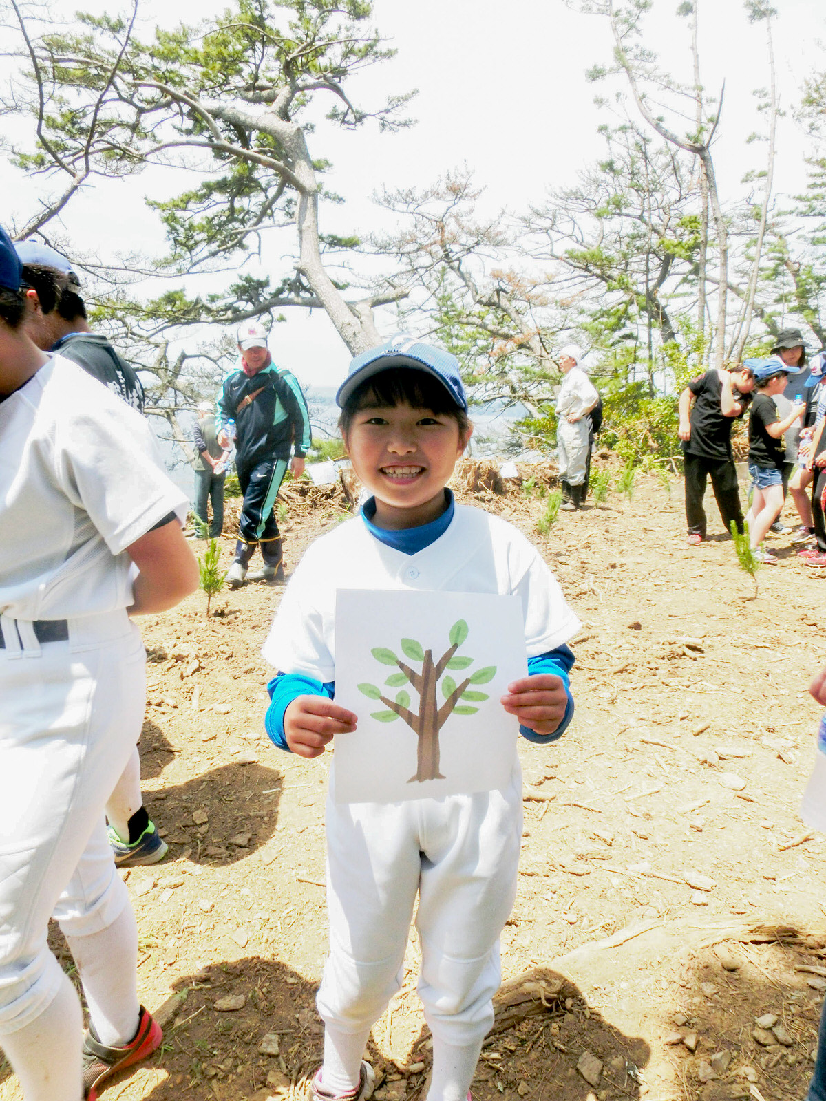 植樹に参加した人々の中、野球のユニフォームを着た女の子が木のイラストが描かれた白い紙を持って笑顔でカメラを見ている写真