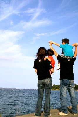 青空と水面を背景に、両親が二人のお子さんを抱いたり肩車したりして、岸壁のような場所に立って海を眺めている様子の写真